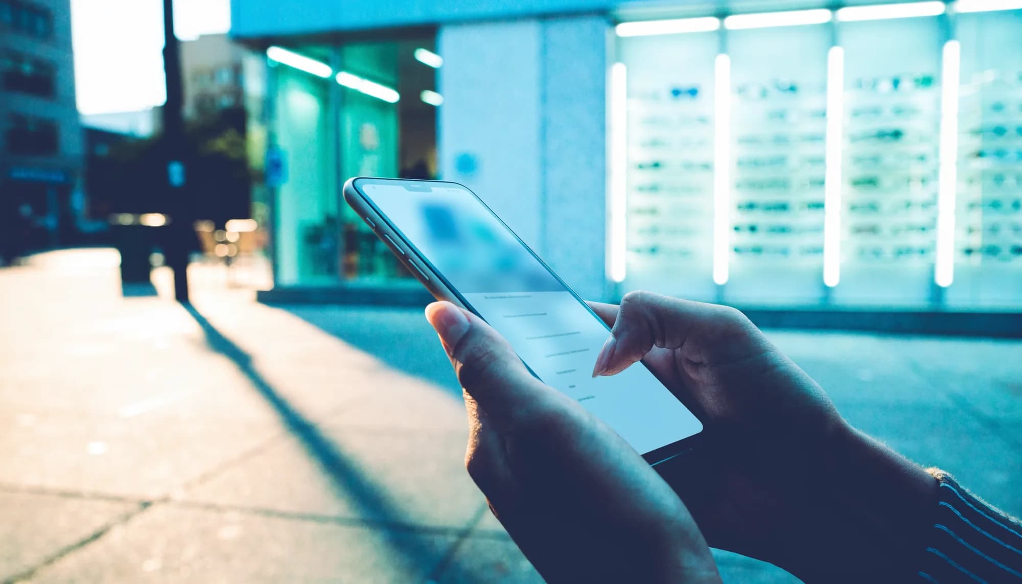 Hand mit einem Smartphone vor dem Schaufenster eines modernen Optikergeschäfts bei Abendlicht.