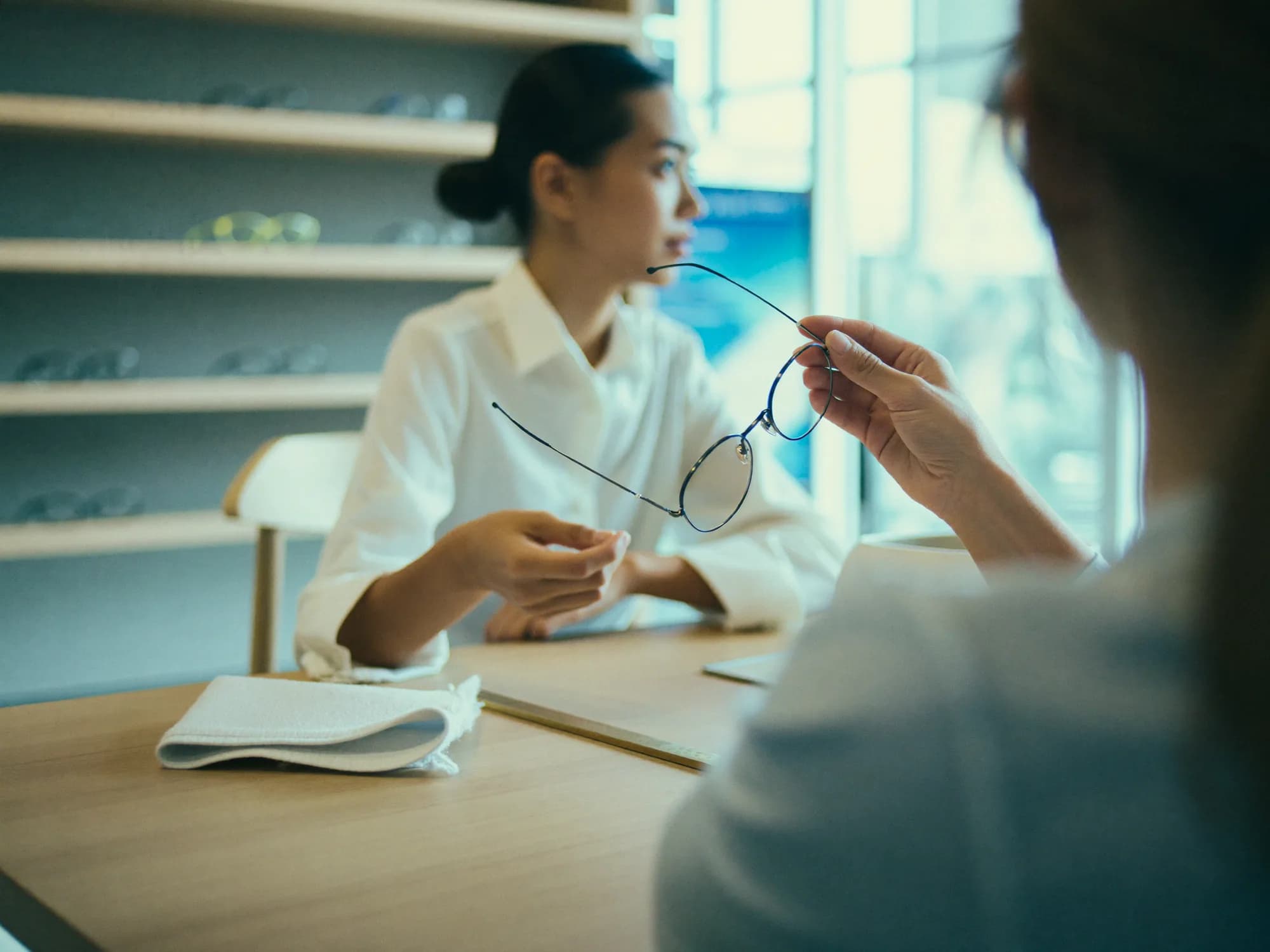 Beratung in der Augenoptik – die Grundlage, die eine Website online fortsetzen muss Kundin hält am Beratungstisch eines Optikergeschäfts eine Brillenfassung hoch, gegenüber sitzt die Augenoptikerin, im Hintergrund eine Wand mit Fassungen